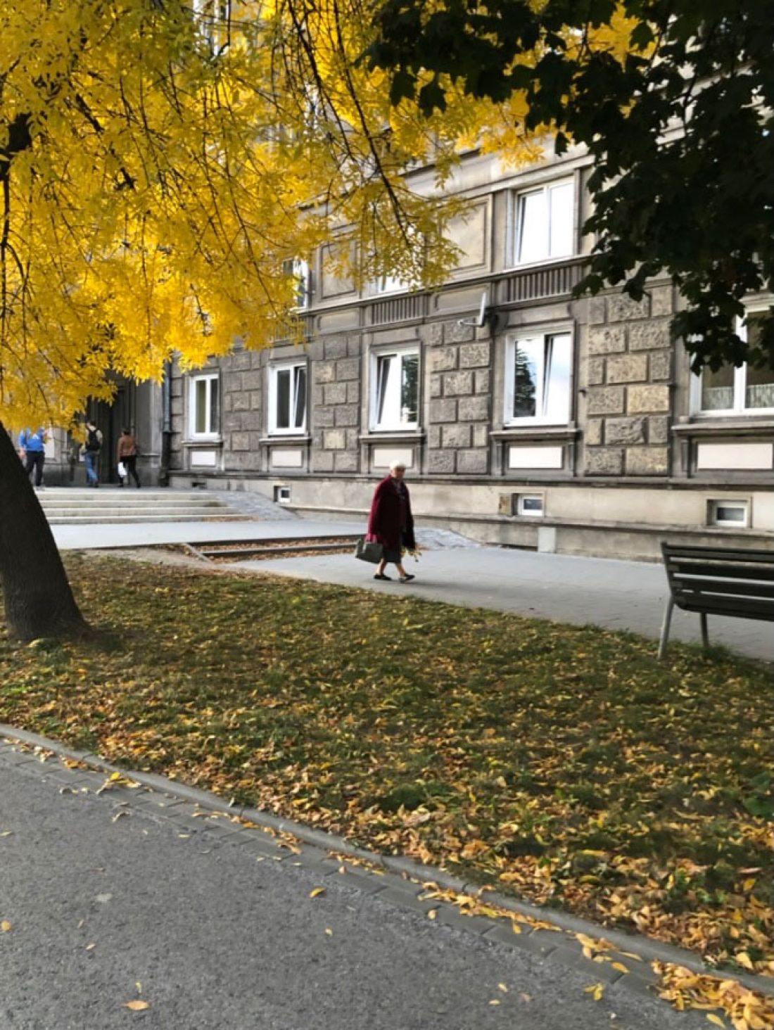 Zdjęcie szerokiego chodnika, odseparowane pasem zieleni od ulicy, w rogu ławka - miejsce do odpoczywania. 