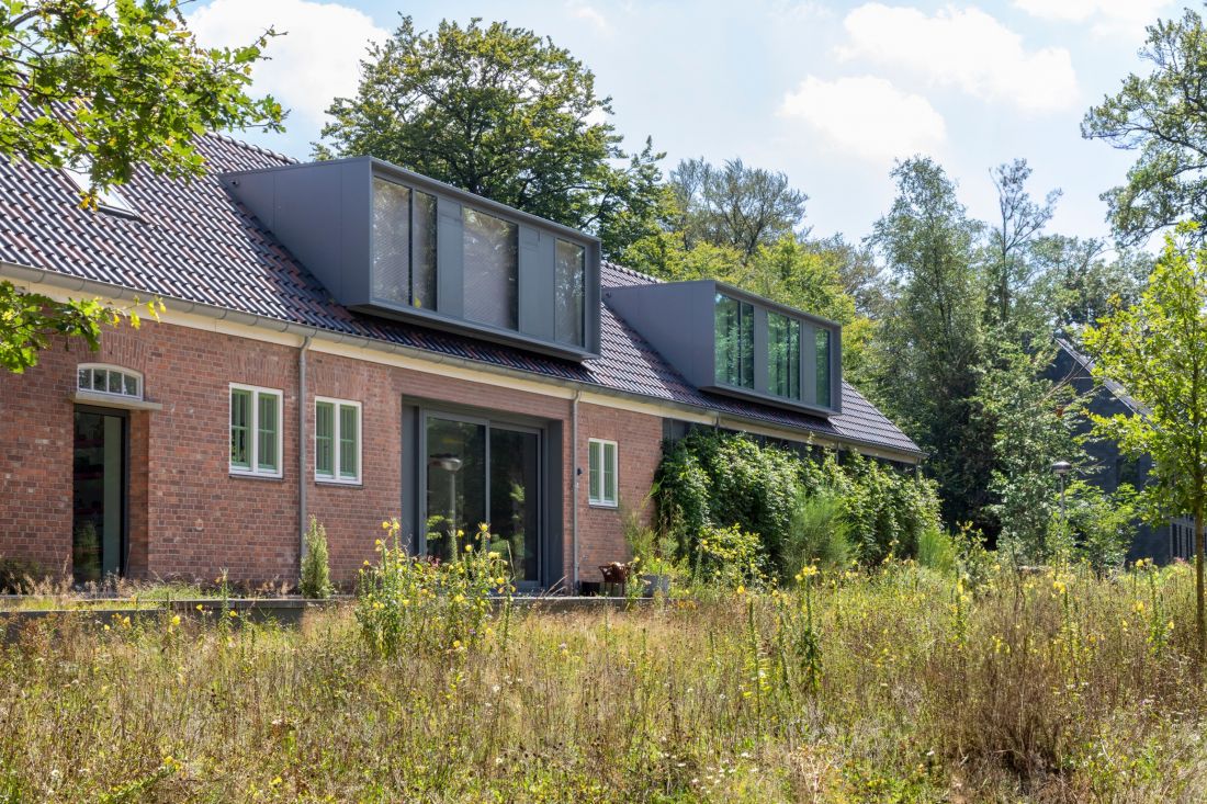 Odrestaurowany dom z czerwonej cegły w lesie, z wieloma oknami, ciemnym dachem i nowoczesnymi elementami architektonicznymi, otoczony dziką trawą oraz polnymi kwiatami