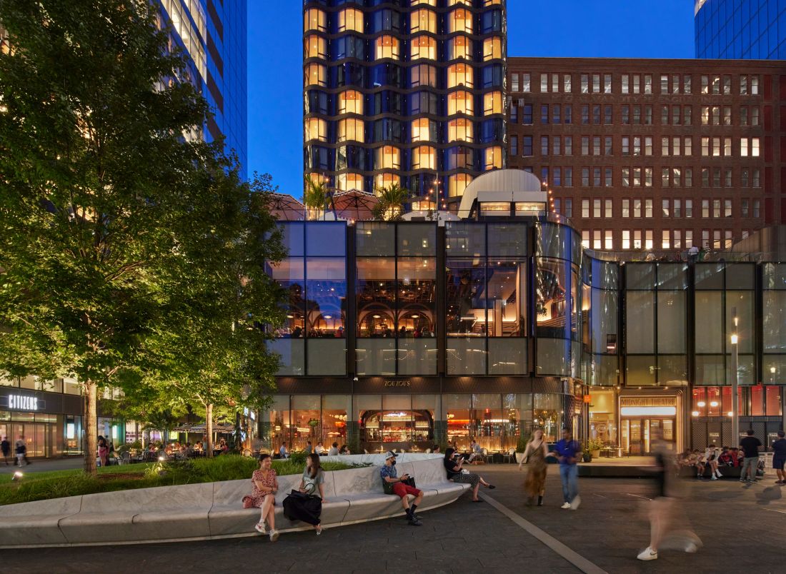 Miejski plac przy Manhattan West o zmierzchu, w tle nowoczesne budynki o szklanych ścianach i ludzie siedzący na zakrzywionej, designerskiej ławce