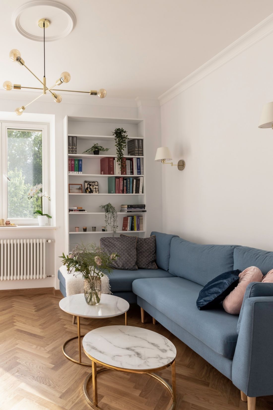 W salonie znajduje się granatowa sofa, nad którą zawisła nowoczesna lampa o kilku żarówkach. Tuż przed nią swoją dwa stoliki o marmurowych blatach 