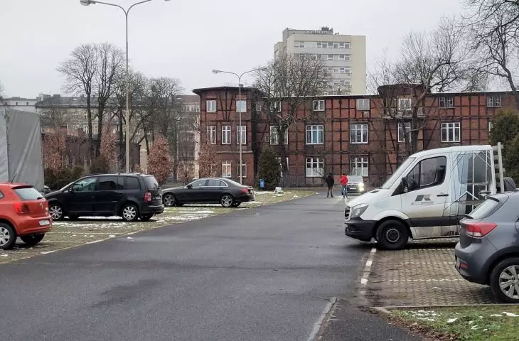 Teren dawnych koszar, parking i widok na budynek szwadronowy, w tle wieżowiec Spóldzielni Mieszkaniowej Grunwald