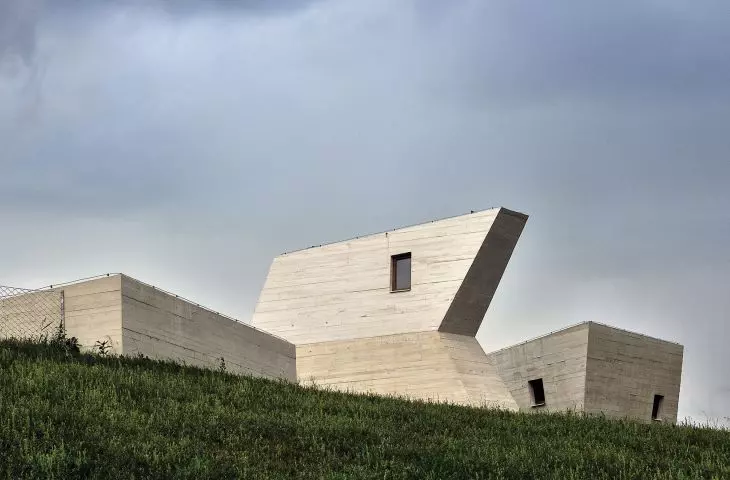 Geometrized buildings contrast with green, slightly undulating terrain; close-up view shows reflections of formwork on raw concrete facade