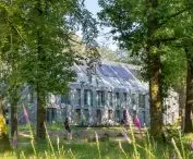 A modern building with large windows and solar panels on the roof, surrounded by lush greenery and tall trees, with purple flowers in the foreground and a person riding on the bike path