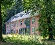 A restored red brick house in the woods, with many windows and a dark roof, surrounded by large deciduous trees