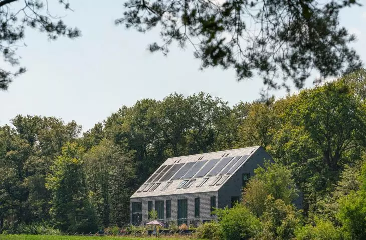 Grey house in the forest with many windows, dark roof and solar panels, surrounded by tall trees and garden