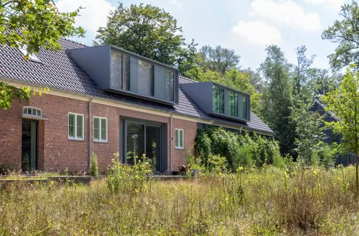 A restored red brick house in the woods, with many windows, a dark roof and modern architectural elements, surrounded by wild grass and field flowers