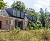 Odrestaurowany dom z czerwonej cegły w lesie, z wieloma oknami, ciemnym dachem i nowoczesnymi elementami architektonicznymi, otoczony dziką trawą oraz polnymi kwiatami