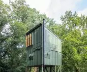 Minimalist wooden building built on an elevation with a green facade, located in a lush forest, with seating areas on a wooden terrace
