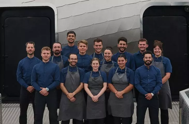 Grupa pracowników restauracji w niebieskich koszulach i szarych fartuchach stojących przed nowoczesnym budynkiem z kopułą i ścianami z wzorem rybiej łuski