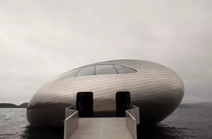 A metal building with an elliptical shape, with two entrances, over the water connected to a pier, surrounded by an overcast sky