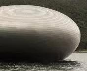 Oval-shaped restaurant building on the water with a wooded hill in the background