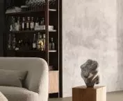 Nowoczesny salon z beżowym fotelem, abstrakcyjną rzeźbą na drewnianym postumencie oraz drewnianym regałem na wino