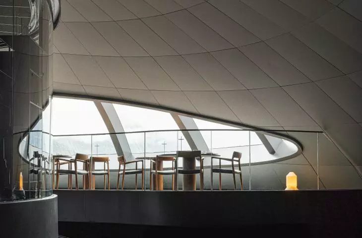 Nowoczesne wnętrze z antresolą, szklaną balustradą, drewnianymi krzesłami i stołami, dużymi oknami i geometrycznym sufitem wyłożonym jasnymi panelami