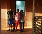 Four boys stand in front of a wall in shades of orange and brown , textured glass window in the background
