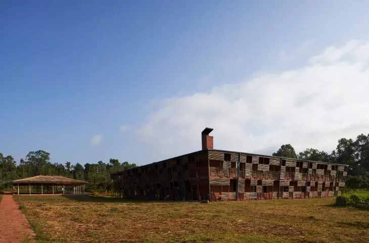 A large rustic building with a slatted facade and chimney, next to a thatched pavilion, set on a dry grassy area under a blue sky