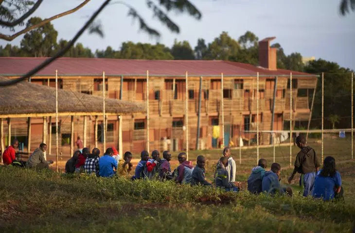 Children sitting on the grass with the large wooden Busajo Campus building in the background