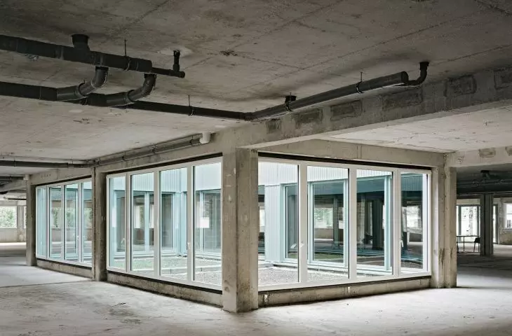 Wnętrze niedokończonego betonowego budynku z centralnym przeszklonym pomieszczeniem i widocznymi rurami sufitowymi