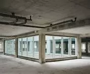 Interior of unfinished concrete building with central glass room and visible ceiling pipes