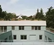 Nowoczesny zielony budynek z fasadą wykończoną blachą falistą i prostokątnymi oknami, na tle drzew i sąsiadujących budynków