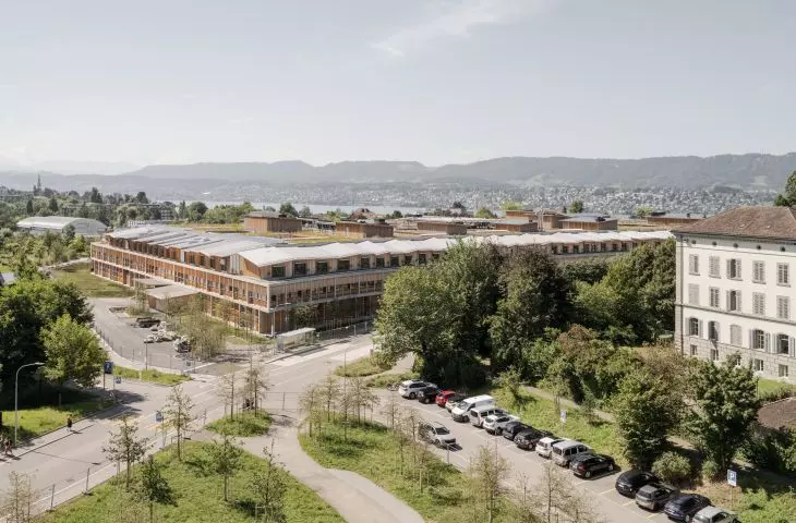 Modern building complex with wooden facades and green roofs surrounded by roads and trees, with the city skyline and mountains in the background