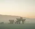 Desert scene at sunset with wooden structures, silhouettes of figures and parked bicycles surrounded by mist