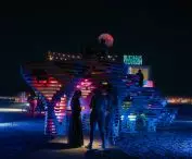 Colorful, illuminated installation at night with people standing around it and a neon sign