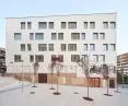 Modern five-story building with white walls, surrounded by a paved courtyard with young trees and street lamps