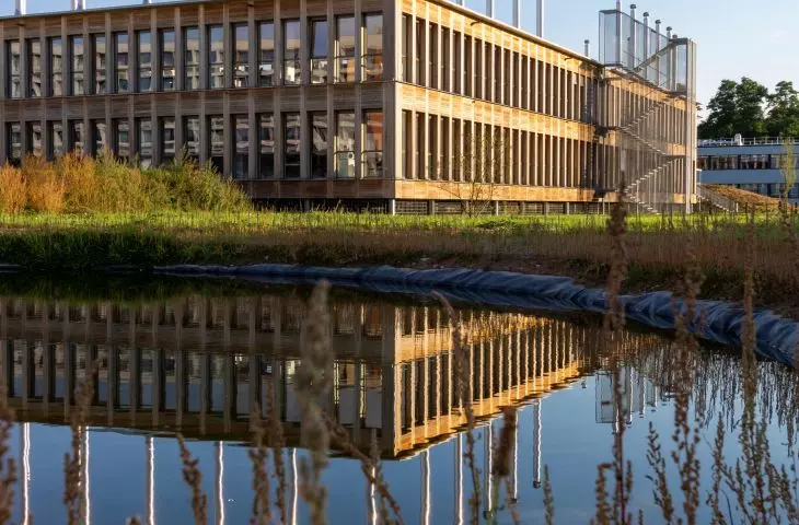 Modern rectangular building with vertical glass panels reflecting in a nearby pond, surrounded by tall grass