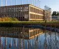 Modern rectangular building with vertical glass panels reflecting in a nearby pond, surrounded by tall grass