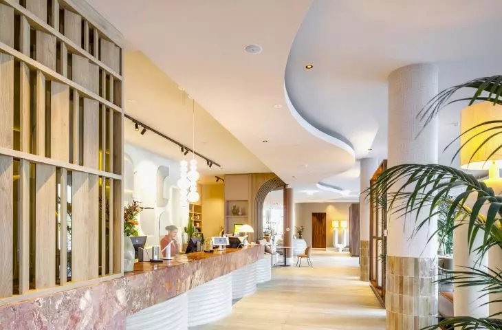 The undulating shapes of Eleganza acoustic ceilings by Knauf Ceiling Solutions harmonize with the shapes of the reception area, while plush, soft sofas in gold and pink colors enhance the effect of friendly acoustics and relaxation 