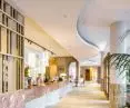 The undulating shapes of Eleganza acoustic ceilings by Knauf Ceiling Solutions harmonize with the shapes of the reception area, while plush, soft sofas in gold and pink colors enhance the effect of friendly acoustics and relaxation 