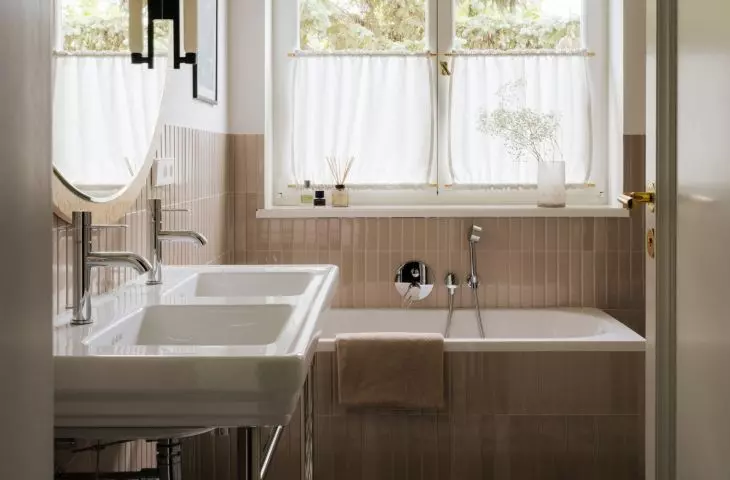 The bright bathroom with a large window has a retro feel. The checkerboard pattern on the floor and the soft curtains add to its homey charm.