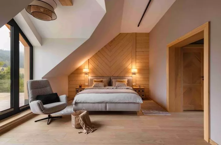 The bedroom with sloped ceilings features a large window offering a view of the Beskid landscape, and the wooden wall behind the bed emphasizes the home’s mountain character