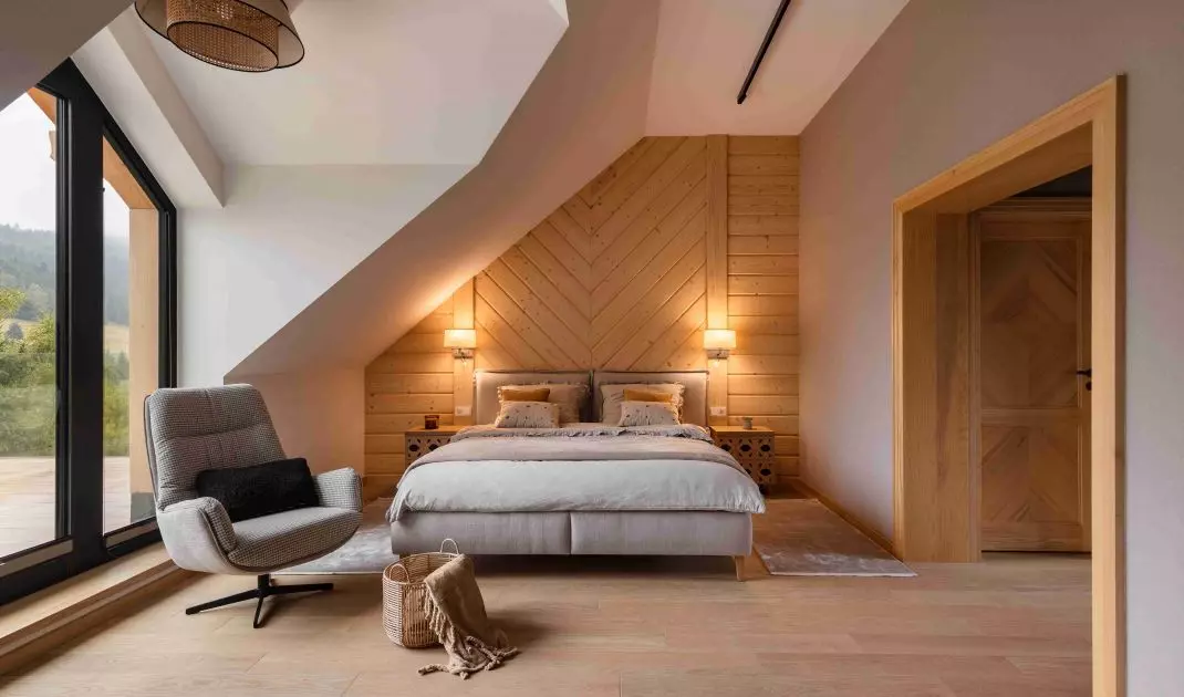 The bedroom with sloped ceilings features a large window offering a view of the Beskid landscape, and the wooden wall behind the bed emphasizes the home’s mountain character