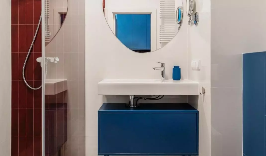 Kolorystyka apartamentu powraca w łazience za sprawą burgundowych płytek i niebieskiej szafki pod umywalką
