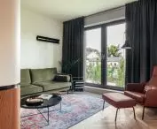 zielona, sztruksowa sofa, skórzany fotel, kolorowy dywan i stylowy gazetnik z lat 70. urozmaicają salon